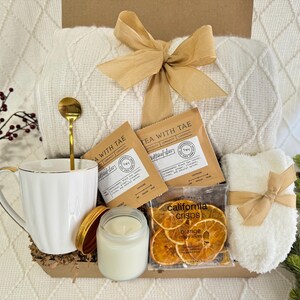 Peut inclure: Un panier-cadeau rempli d'articles confortables pour une soir&eacute;e relaxante. Il comprend une couverture en tricot blanc, une tasse blanche, une cuill&egrave;re dor&eacute;e, une bougie, une bo&icirc;te de chips d'orange et deux sachets de th&eacute; &eacute;tiquet&eacute;s "Tea with Tae".