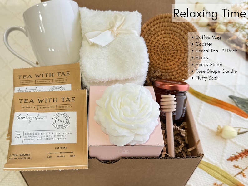May include: A gift box filled with relaxing items, including a white coffee mug, two tea sachets labeled "TEA WITH TAE", a white fluffy sock, a white rose-shaped candle, a small jar of honey with a wooden honey dipper, a white fluffy towel, and a woven coaster.