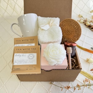 Peut inclure: Une bo&icirc;te-cadeau en carton marron remplie d'une tasse blanche, une serviette blanche, une fleur blanche, des sachets de th&eacute;, un pot de miel, un b&acirc;tonnet de miel en bois et un dessous-de-verre tiss&eacute;.
