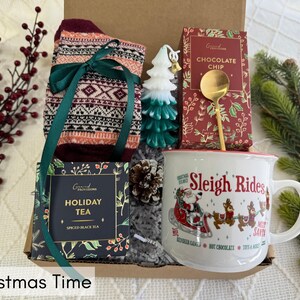 May include: A Christmas gift basket with a mug that says "Sleigh Rides", a box of holiday tea, a pair of patterned socks, a small tree ornament, and a box of chocolate chips.