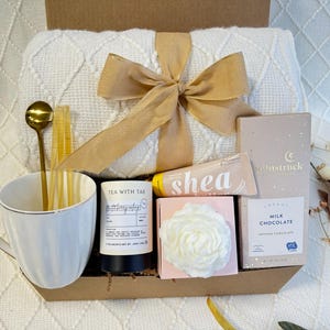 May include: A gift basket filled with self-care items, including a white mug, a white throw blanket, a bar of soap, a candle, and a box of chocolate. The basket is tied with a brown ribbon.
