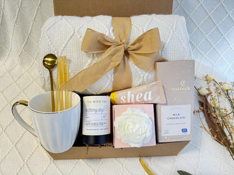 May include: A gift basket filled with self-care items, including a white mug, a white throw blanket, a bar of soap, a candle, and a box of chocolate. The basket is tied with a brown ribbon.