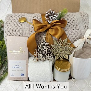 Puede incluir: Una caja de regalo con un tema acogedor, que presenta una manta de punto beige, una cuchara dorada, una barra de chocolate, una taza blanca, una vela y un copo de nieve decorativo. La caja est&aacute; atada con una cinta dorada e incluye un letrero de madera que dice "A very Merry Holly Jolly Christmas". El texto "All I Want is You" est&aacute; en la parte inferior.
