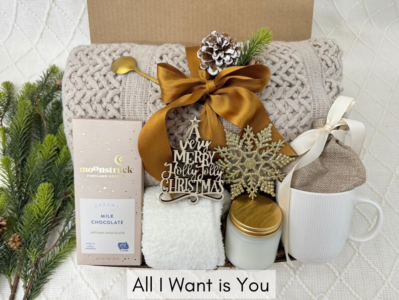 Puede incluir: Una caja de regalo con un tema acogedor, que presenta una manta de punto beige, una cuchara dorada, una barra de chocolate, una taza blanca, una vela y un copo de nieve decorativo. La caja est&aacute; atada con una cinta dorada e incluye un letrero de madera que dice "A very Merry Holly Jolly Christmas". El texto "All I Want is You" est&aacute; en la parte inferior.