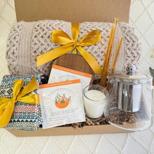 May include: A gift basket filled with cozy items, including a knitted throw blanket, a patterned scarf, a glass tea pot with a stainless steel infuser, a candle, and two boxes of lavender orange grey tea.