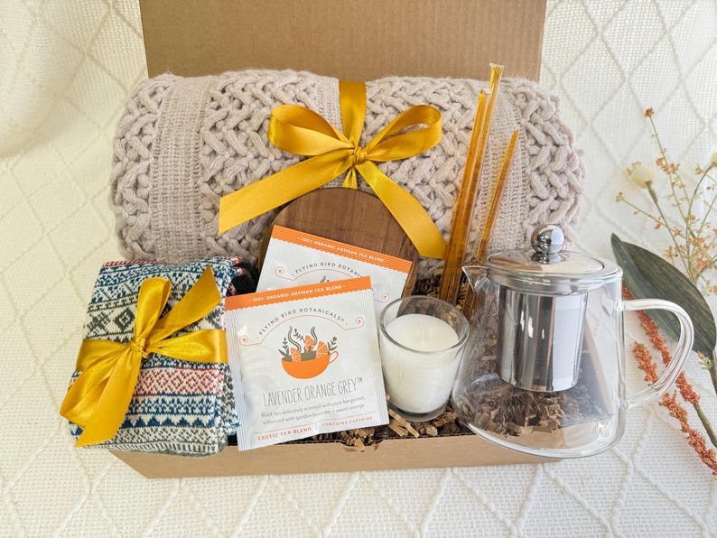 May include: A gift basket filled with cozy items, including a knitted throw blanket, a patterned scarf, a glass tea pot with a stainless steel infuser, a candle, and two boxes of lavender orange grey tea.