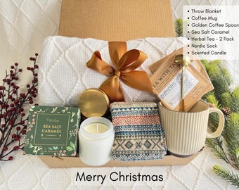 Può includere: Un cesto regalo pieno di articoli per una confortevole serata invernale. Il cesto include una coperta, una tazza da caffè, una candela, una bustina di tè, un cucchiaio dorato e un paio di calzini. Il cesto è decorato con un nastro marrone e un rametto di bacche rosse.