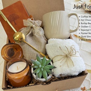 May include: A gift box filled with items for self-care and relaxation. The box includes a coffee mug, a candle, a small succulent, a notebook, a pen, a burlap bag, and a towel.