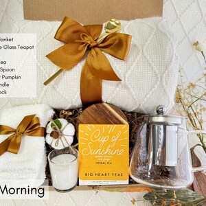 May include: A Fall Morning gift basket with a white throw blanket, a glass teapot, a candle, a hand towel, a mini knit pumpkin, a golden spoon, and a box of herbal tea labeled "Cup of Sunshine".