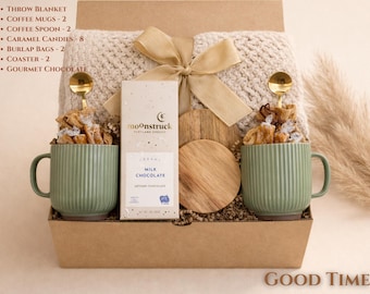 May include: A gift basket with a cream-colored throw blanket, two green ribbed coffee mugs, caramel candies, and a bar of milk chocolate. The basket includes two gold coffee spoons, burlap bags, and coasters. The words "GOOD TIME" are at the bottom.