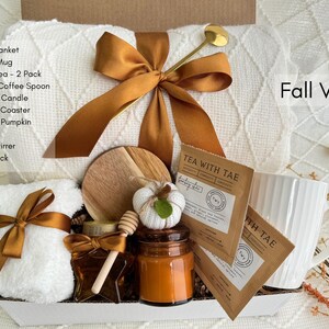 May include: A white gift box filled with fall-themed items, including a throw blanket, coffee mug, tea, candle, and a mini knit pumpkin. The box is tied with a gold ribbon and the words "Fall Vibes" are printed on the side.