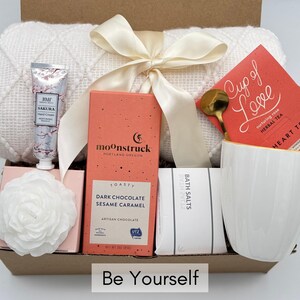 May include: A gift box filled with self-care items, including a white throw blanket, a white mug, a bar of chocolate, bath salts, a hand cream, and a box of herbal tea. The box is decorated with a white ribbon and the words "Be Yourself".