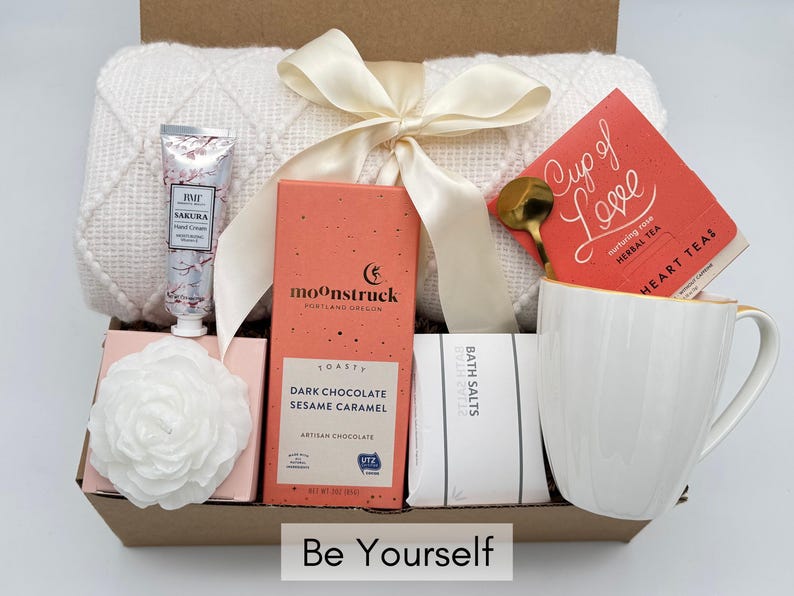 May include: A gift box filled with self-care items, including a white throw blanket, a white mug, a bar of chocolate, bath salts, a hand cream, and a box of herbal tea. The box is decorated with a white ribbon and the words "Be Yourself".