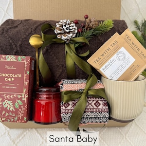 Puede incluir: Una caja de regalo con una manta marr&oacute;n, galletas con chispas de chocolate, una vela roja, calcetines estampados, una taza con bolsitas de t&eacute; y una cuchara dorada. La caja est&aacute; decorada con cintas verdes y pi&ntilde;as. El texto en la caja dice "Santa Baby."