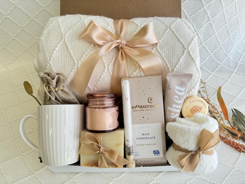 Peut inclure: Un panier-cadeau blanc rempli d'articles de soins personnels, dont une couverture blanche, une tasse, une bougie, du savon, des produits de bain et du chocolat. Le panier est attach&eacute; avec un ruban beige.