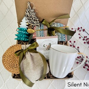 May include: A gift box filled with items for a cozy night in, including a mug, a bag of tea, a wooden ornament, and a woven coaster. The box is decorated with a green and red plaid fabric and the text "Silent Night".