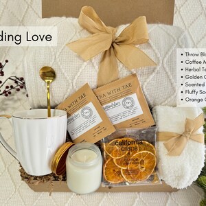 K&ouml;nnte beinhalten: Ein Geschenk-Korb mit einer wei&szlig;en Tasse, einem goldenen L&ouml;ffel, Kr&auml;utertee, einer Kerze, flauschigen Socken und knusprigen Orangenscheiben. Der Korb enth&auml;lt eine Decke und ist mit einem goldenen Band gebunden. Der Text "Sending Love" ist sichtbar.