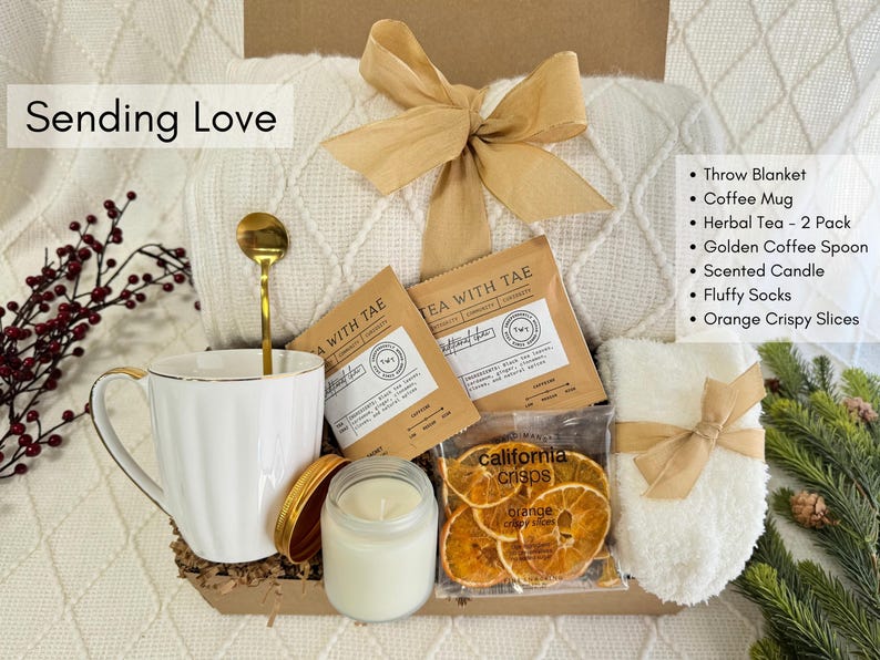 K&ouml;nnte beinhalten: Ein Geschenk-Korb mit einer wei&szlig;en Tasse, einem goldenen L&ouml;ffel, Kr&auml;utertee, einer Kerze, flauschigen Socken und knusprigen Orangenscheiben. Der Korb enth&auml;lt eine Decke und ist mit einem goldenen Band gebunden. Der Text "Sending Love" ist sichtbar.