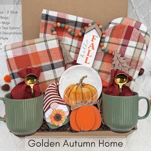 K&ouml;nnte beinhalten: Ein herbstliches Geschenkset mit zwei gr&uuml;nen Kaffeetassen, einem kleinen wei&szlig;en Teller mit einem K&uuml;rbis und herbstlichen Dekorationen. Enth&auml;lt einen Mini-Teller, Servietten und einen Ofenhandschuh. Der Text "Golden Autumn Home" steht unten.