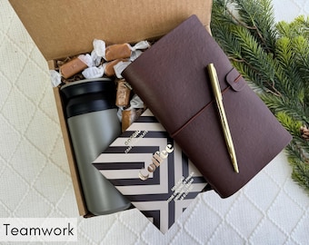 May include: A gift box containing a gray travel mug, a brown leather journal with a gold pen, a chocolate bar, and wrapped candies. The box is lined with brown paper and sits on a white patterned surface. The word "Teamwork" is at the bottom.