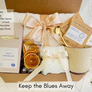 May include: A gift basket filled with items for relaxation and comfort. It includes a throw blanket, a mug, a tea bag, a bar of chocolate, orange slices, and a golden spoon. The basket is decorated with a beige ribbon and a white towel. The text "Keep the Blues Away" is printed on the bottom of the basket.
