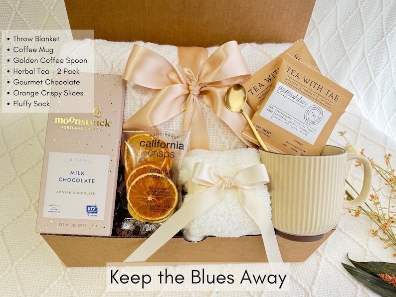 May include: A gift basket filled with items for relaxation and comfort. It includes a throw blanket, a mug, a tea bag, a bar of chocolate, orange slices, and a golden spoon. The basket is decorated with a beige ribbon and a white towel. The text "Keep the Blues Away" is printed on the bottom of the basket.