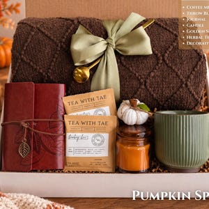 May include: A gift box containing a dark brown knit throw blanket with a sage green ribbon and gold spoon, a red leather journal with a leaf charm, two Tea With Tae herbal tea sachets, a small candle, and a decorative white pumpkin mug.
