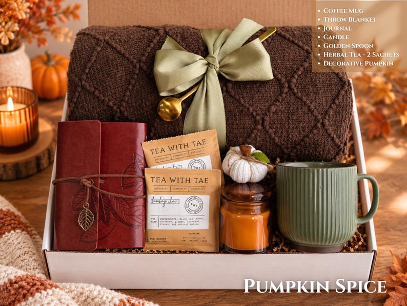 May include: A gift box containing a dark brown knit throw blanket with a sage green ribbon and gold spoon, a red leather journal with a leaf charm, two Tea With Tae herbal tea sachets, a small candle, and a decorative white pumpkin mug.