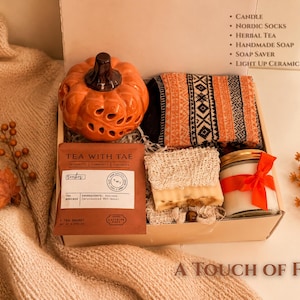 May include: A gift box containing a ceramic orange pumpkin, patterned socks, a tea sachet labeled Tea With Tae, a bar of soap in a mesh soap saver, and a candle with a red ribbon. The box lid lists the contents: Candle, Nordic Socks, Herbal Tea, Handmade Soap, Soap Saver, Light Up Ceramic Pumpkin.