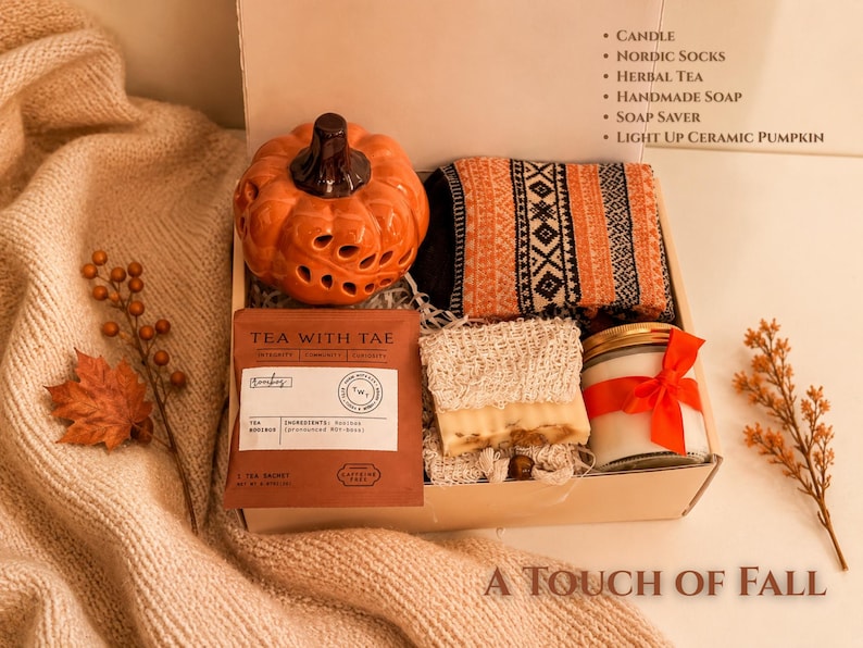 May include: A gift box containing a ceramic orange pumpkin, patterned socks, a tea sachet labeled Tea With Tae, a bar of soap in a mesh soap saver, and a candle with a red ribbon. The box lid lists the contents: Candle, Nordic Socks, Herbal Tea, Handmade Soap, Soap Saver, Light Up Ceramic Pumpkin.