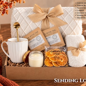 May include: A gift box containing a white ribbed coffee mug with a golden spoon, a cream-colored throw blanket with a tan ribbon, a pair of white fluffy socks, a small candle, two Tea With Tae herbal tea sachets, and a bag of California Crisps dried orange slices.