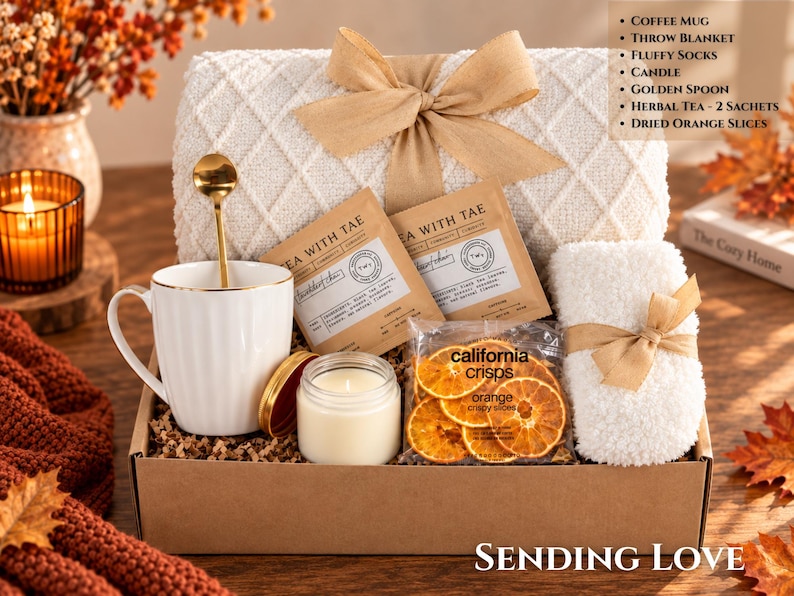 May include: A gift box containing a white ribbed coffee mug with a golden spoon, a cream-colored throw blanket with a tan ribbon, a pair of white fluffy socks, a small candle, two Tea With Tae herbal tea sachets, and a bag of California Crisps dried orange slices.