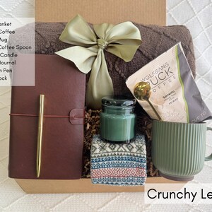 K&ouml;nnte beinhalten: Eine Geschenkbox mit einer braunen Decke, Kaffee, einer gr&uuml;nen Tasse, einem goldenen L&ouml;ffel, einer Duftkerze, einem Notizbuch, einem goldenen Stift und einer nordischen Socke. Die Box ist mit einer gr&uuml;nen Schleife gebunden. Der Text "Crunchy Leaves" steht unten.