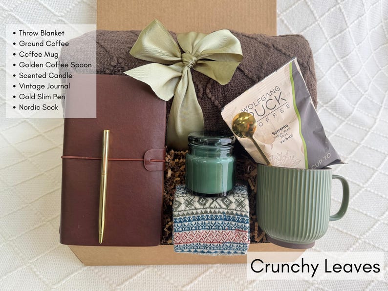 K&ouml;nnte beinhalten: Eine Geschenkbox mit einer braunen Decke, Kaffee, einer gr&uuml;nen Tasse, einem goldenen L&ouml;ffel, einer Duftkerze, einem Notizbuch, einem goldenen Stift und einer nordischen Socke. Die Box ist mit einer gr&uuml;nen Schleife gebunden. Der Text "Crunchy Leaves" steht unten.