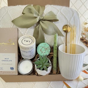 May include: A gift basket filled with self-care items, including a white blanket, a mug, a candle, a small succulent plant, a bar of milk chocolate, a tea bag, and bath salts. The basket is lined with brown paper and green shredded paper.