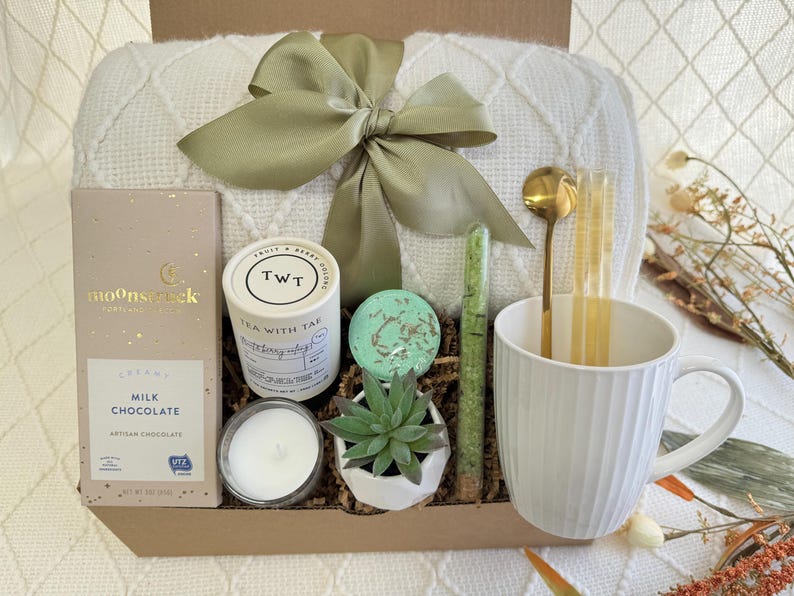 May include: A gift basket filled with self-care items, including a white blanket, a mug, a candle, a small succulent plant, a bar of milk chocolate, a tea bag, and bath salts. The basket is lined with brown paper and green shredded paper.