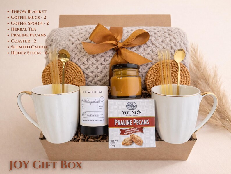 May include: A gift box with a gray throw blanket, two white coffee mugs with gold trim, and a jar of honey. The box also contains a candle, tea, praline pecans, and honey sticks. The text "JOY GIFT BOX" is at the bottom.