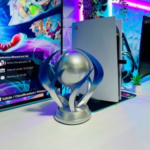 May include: A silver trophy with a blue and white design, featuring the PlayStation logo on a sphere. The trophy is sitting on a white surface in front of a white PlayStation 5 console.