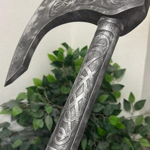 May include: A silver-coloured, curved axe with intricate Celtic knotwork engravings on the blade and handle. The handle is a dark brown colour. The axe is a prop or decorative item, possibly for cosplay or historical reenactment.