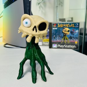 Puede incluir: Figura impresa en 3D de un personaje esquelético con una base verde. El cráneo es beige con un ojo azul y una cuenca negra. La base es una estructura verde en forma de mano. Una caja de juego de PlayStation 2 es visible en el fondo.