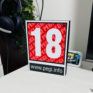 May include: A black and white sign with a red background. The sign has the number 18 in white and the text "www.pegi.info" in black. The background is a repeating pattern of the word "PEGI" in red.