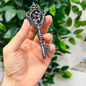Pode incluir: Chave prateada ornamentada com um design decorativo de querubim no topo. A chave tem uma al&ccedil;a torcida e um detalhe da chave. O fundo est&aacute; desfocado com folhagem verde.