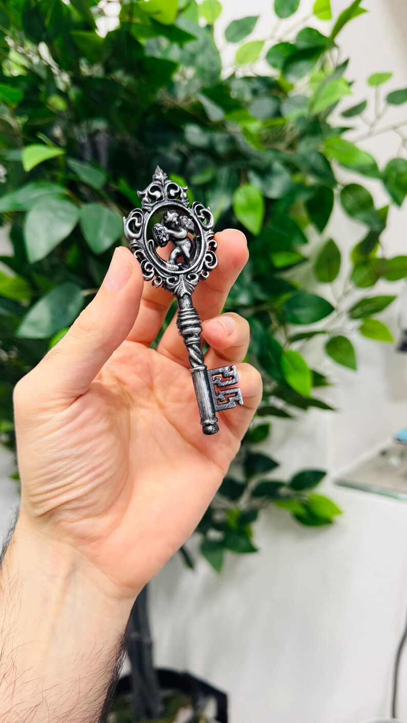 Pode incluir: Chave prateada ornamentada com um design decorativo de querubim no topo. A chave tem uma al&ccedil;a torcida e um detalhe da chave. O fundo est&aacute; desfocado com folhagem verde.