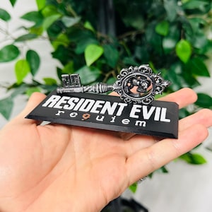 Pode incluir: Uma chave decorativa com o texto "RESIDENT EVIL requiem" em uma base preta. A chave &eacute; prateada com detalhes ornamentados, incluindo um emblema circular. O fundo apresenta folhagem verde.