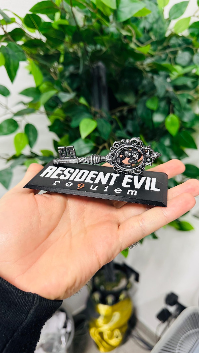 Pode incluir: Uma chave decorativa com o texto "RESIDENT EVIL requiem" em uma base preta. A chave &eacute; prateada com detalhes ornamentados, incluindo um emblema circular. O fundo apresenta folhagem verde.