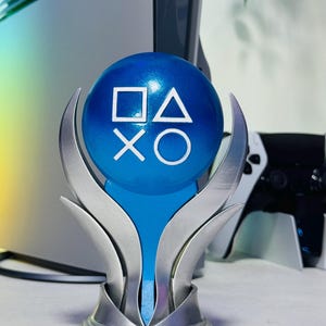Pode incluir: Um troféu esférico azul com os símbolos brancos do comando da PlayStation: quadrado, triângulo, cruz e círculo. A esfera é mantida por uma base metálica prateada de várias pontas que se curva para cima para apoiar o orbe em exibição.