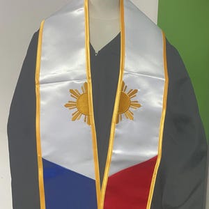 May include: A white graduation sash with gold trim and a blue and red section. The sash features a gold embroidered sun with rays, a symbol of the Philippine flag.