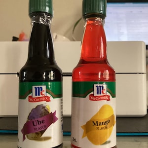 Könnte beinhalten: Zwei kleine Glasflaschen mit grünen Deckeln und weißen Etiketten. Die Flaschen enthalten McCormick Ube Flavor und McCormick Mango Flavor. Die Etiketten zeigen Illustrationen der Früchte.