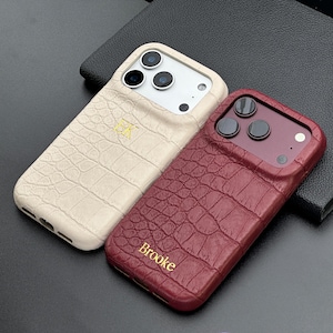May include: Two smartphone cases with a textured crocodile skin pattern. One case is cream-colored with the gold embossed letters EK, and the other is deep red with the gold embossed name Brooke. Both feature cutouts for camera lenses.
