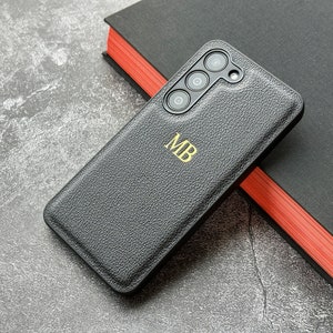 May include: A black leather phone case with a gold monogram "MB" and a camera lens cutout.